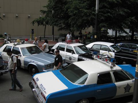 1991 Ford LTD Crown Victoria - cluster of pale-blue-and-white POLICE cars with star emblems and roof beacons are parked together as people walk among them.