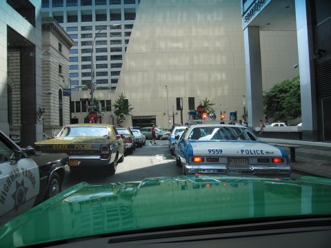 1991 Ford LTD Crown Victoria - View over a green hood at a lineup of vintage police cruisers staged in a downtown street between office towers: a blue-and-white 1970s Che…