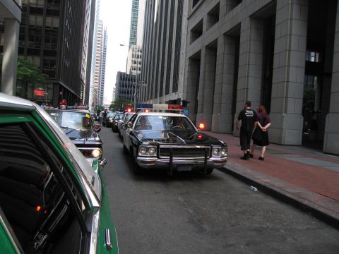 1991 Ford LTD Crown Victoria - black vintage police sedan with red roof lights lit drives down a narrow street flanked by tall office towers.