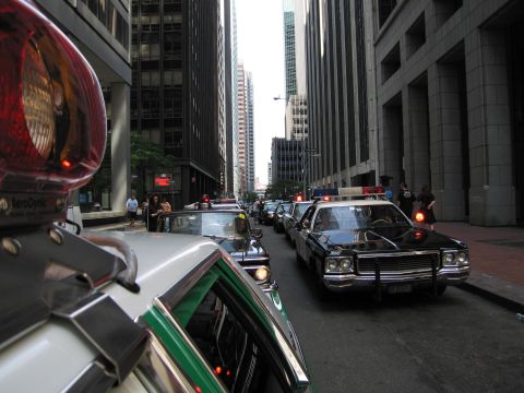 1991 Ford LTD Crown Victoria - convoy of classic police cars with lit roof beacons proceeds down a narrow street between tall buildings.