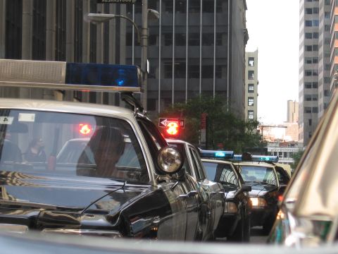 1991 Ford LTD Crown Victoria - Vintage police cars with red and blue roof beacons sit in a line at a downtown intersection under a red traffic signal.