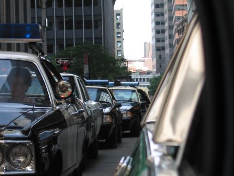 1991 Ford LTD Crown Victoria - line of black classic police cars with chrome spotlights and roof beacons stretches down a city street toward distant buildings.
