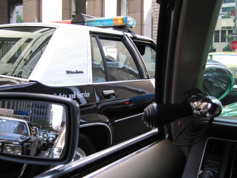 1991 Ford LTD Crown Victoria - black-and-white police car marked Safety and Service with a colorful lightbar, viewed through the window and mirror of another car.