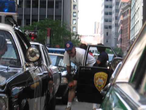 1991 Ford LTD Crown Victoria - People gather between squad cars on a city street, one car bearing a yellow sheriff-style department emblem on its door.