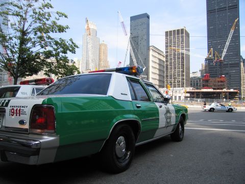 1991 Ford LTD Crown Victoria - green-and-white sheriff cruiser with EMERGENCY 911 markings and a star emblem sits before a skyline with tower cranes.