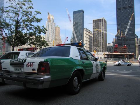 1991 Ford LTD Crown Victoria - green-and-white sheriff sedan with 911 emergency markings is parked on a street with cranes and skyscrapers behind it.