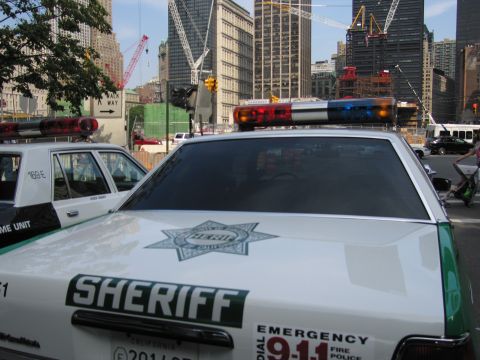 1991 Ford LTD Crown Victoria - rear quarter of a white sheriff car shows a green star emblem and EMERGENCY 911 markings before a crane-filled skyline.