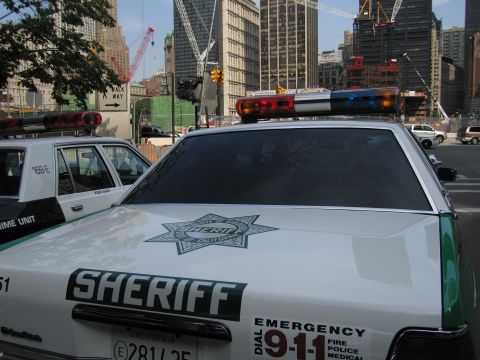 1991 Ford LTD Crown Victoria - white sheriff car with a green star emblem, lightbar and 911 markings stands before downtown towers under construction.
