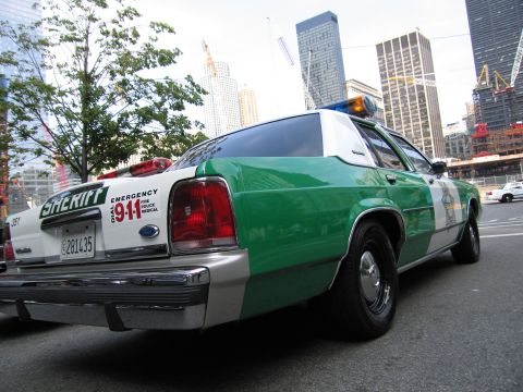 1991 Ford LTD Crown Victoria - Rear three-quarter view of a green-and-white sheriff sedan with 911 markings parked before a skyline with cranes.