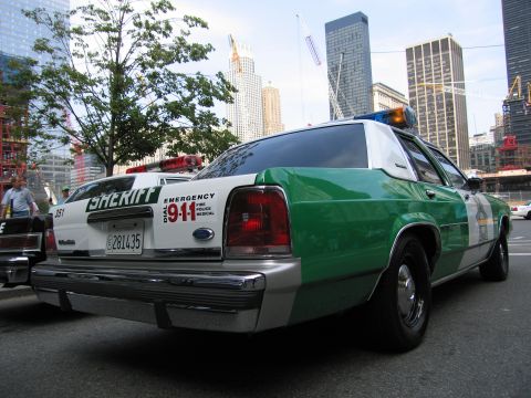 1991 Ford LTD Crown Victoria - green-and-white sheriff sedan with a roof lightbar and 911 markings sits on a street backed by tall city buildings.