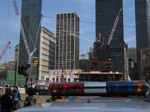 1991 Ford LTD Crown Victoria - police car's red-and-blue lightbar fills the foreground before a downtown skyline dotted with tower cranes and a low billboard.