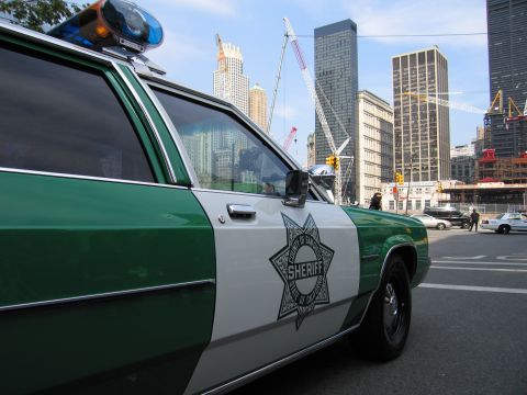 1991 Ford LTD Crown Victoria - Close side view of a green-and-white Sheriff star-badge patrol sedan parked on a city street with high-rise buildings and cranes in the bac…