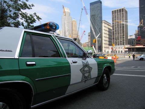 1991 Ford LTD Crown Victoria - green-and-white Sheriff sedan with a star door badge and roof light bar parked at an intersection beside a fenced construction zone with cr…