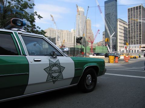 1991 Ford LTD Crown Victoria - Front-quarter view of a green-and-white Sheriff star-badge patrol car parked along a street facing a large fenced construction site with cr…
