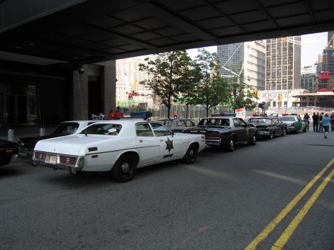 1991 Ford LTD Crown Victoria - white vintage police sedan with a star badge leads a row of classic patrol cars parked under a building overpass on a city street.