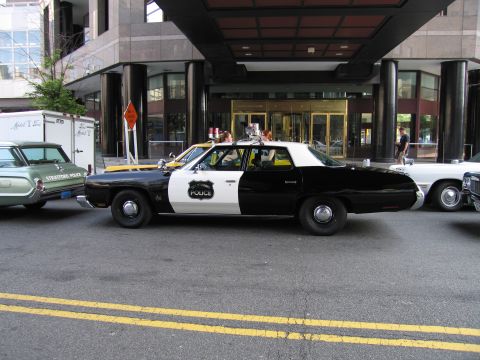 1991 Ford LTD Crown Victoria - black-and-white Police sedan with a roof light bar parked at the curb near a building entrance, flanked by other classic patrol vehicles.