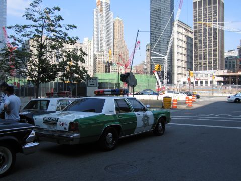1991 Ford LTD Crown Victoria - green-and-white Sheriff patrol sedan with roof lights parked at a city intersection across from a fenced construction site with cranes.