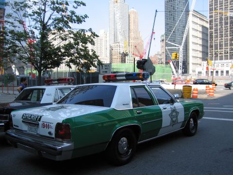 1991 Ford LTD Crown Victoria - Rear-quarter view of a green-and-white Sheriff station-wagon-style patrol car with roof light bar parked at a downtown intersection near cr…