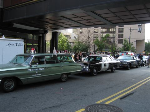 1991 Ford LTD Crown Victoria - green Stratford Police radio-patrol station wagon heads a line of vintage patrol cars parked under a building overpass on a city street.