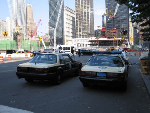 1991 Ford LTD Crown Victoria - Two black-and-tan police sedans parked side by side at a broad city intersection with skyscrapers and construction cranes in the background.