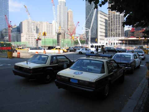 1991 Ford LTD Crown Victoria - Two black-and-tan patrol sedans with door emblems parked at a wide intersection near a fenced construction zone with cranes and high-rise b…