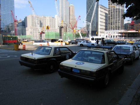 1991 Ford LTD Crown Victoria - pair of black-and-tan patrol sedans with door seals parked at a downtown intersection, with cranes, traffic and skyscrapers behind.