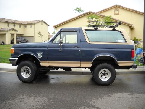 1986 Ford Bronco - This 1987 Ford Bronco features a two-tone paint scheme and a roof rack, parked on a wet street.