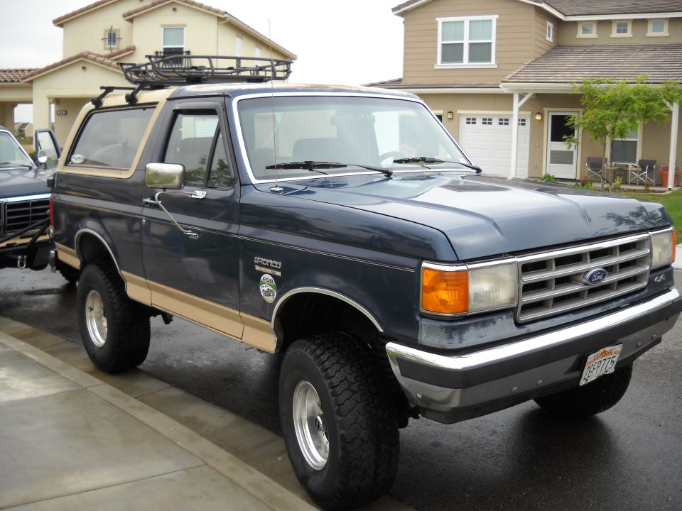 Blue Ford Bronco with tan trim and roof rack parked on street. Another vehicle is partially visible to the left