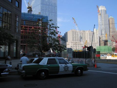 1991 Ford LTD Crown Victoria - green-and-white Sheriff patrol sedan parked at the curb in shadow, facing a steel high-rise under construction with cranes and scaffolding.