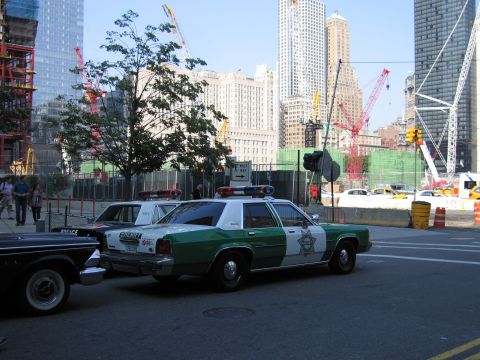 1991 Ford LTD Crown Victoria - Front-quarter view of a green-and-white Sheriff patrol sedan with roof lights parked next to an older black patrol car, facing a constructi…