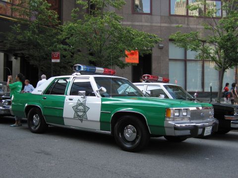 1991 Ford LTD Crown Victoria - green-and-white Sheriff sedan with a six-point star door badge and red-and-blue roof lights parked at the curb beside a building on a city…