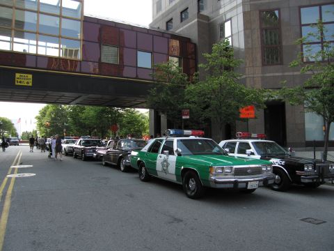 1991 Ford LTD Crown Victoria - row of vintage patrol cars, including a green-and-white Sheriff sedan and a black Police car, parked along a curb beneath a building skybri…