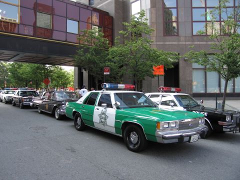 1991 Ford LTD Crown Victoria - green-and-white Sheriff sedan with a star badge heads a line of parked vintage patrol cars along a curb under a building skybridge.