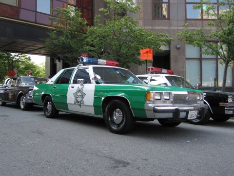 1991 Ford LTD Crown Victoria - Front-quarter view of a green-and-white Sheriff star-badge patrol sedan parked at the curb between other vintage police cars on a city stre…