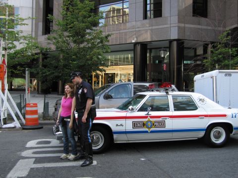 1991 Ford LTD Crown Victoria - uniformed police officer and a woman in pink stand posing beside a white State Police sedan with red-and-blue roof lights on a city street.