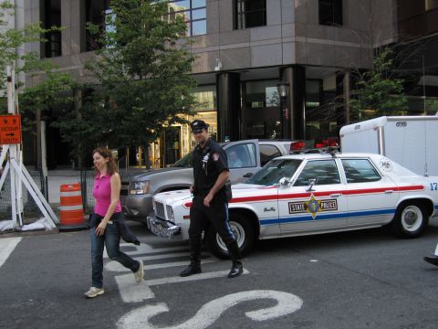 1991 Ford LTD Crown Victoria - uniformed officer and a woman in pink walk across a crosswalk past a white State Police sedan parked on a city street.
