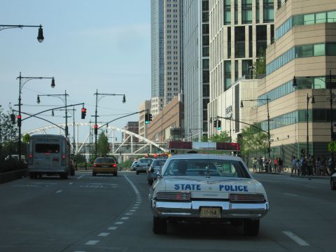 1991 Ford LTD Crown Victoria - vintage white State Police sedan with a red roof lightbar drives down a downtown street past glass office towers and an arched bridge.