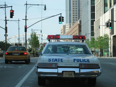 1991 Ford LTD Crown Victoria - Rear view of a white State Police sedan with twin red roof lights, stopped at a green traffic signal beside a yellow taxi downtown.