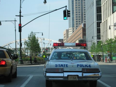 1991 Ford LTD Crown Victoria - white State Police car seen from behind drives a downtown street lined with office buildings and a white arched pedestrian bridge.