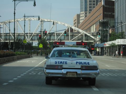 1991 Ford LTD Crown Victoria - white State Police sedan with roof lightbar drives a city street beneath a large white steel truss footbridge.
