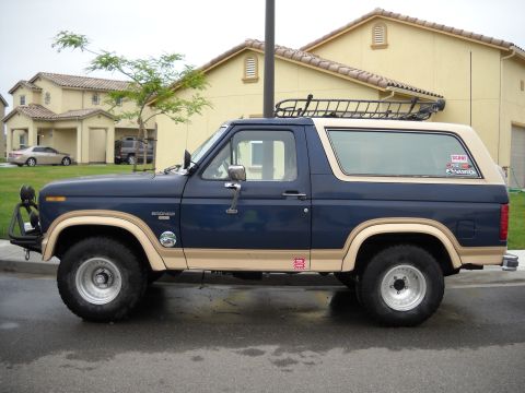 1986 Ford Bronco - This 1986 Ford Bronco features a two-tone paint scheme and a roof rack, seen parked on a wet street.