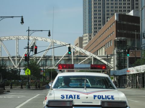1991 Ford LTD Crown Victoria - Close rear view of a white police sedan with its red roof lightbar, approaching traffic signals near a white steel truss footbridge.