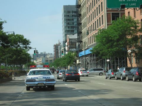 1991 Ford LTD Crown Victoria - white State Police sedan drives a tree-lined city avenue in traffic, with a green highway sign reading New Jersey overhead.