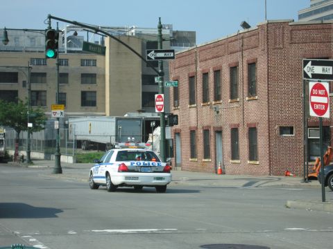 1991 Ford LTD Crown Victoria - white Crown Victoria-style police car turns at a city intersection beside a red brick building with one-way and do-not-enter signs.