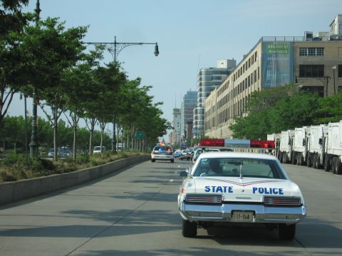 1991 Ford LTD Crown Victoria - Rear view of a white full-size sedan lettered 'STATE POLICE' in blue with a red stripe, running a red roof lightbar down a tree-lined city…