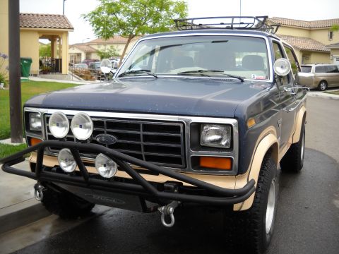 1986 Ford Bronco - A blue Ford Bronco with tan trim and a black bull bar is parked on a wet street.