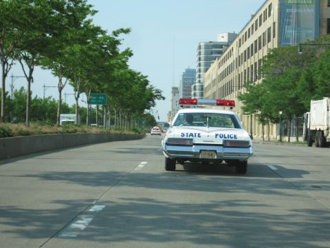 1991 Ford LTD Crown Victoria - Rear view of a white State Police sedan with roof lightbar cruising a divided urban roadway past trees and a parked truck.