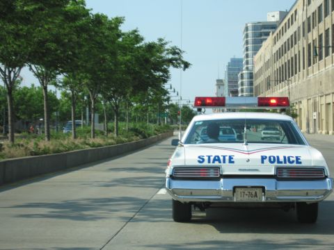 1991 Ford LTD Crown Victoria - white State Police sedan with twin red roof lights drives a curving roadway lined with young trees and tall buildings.