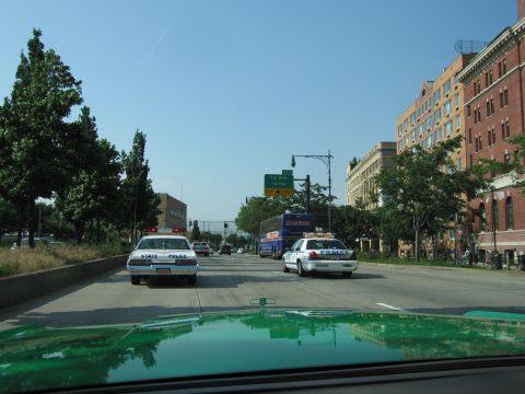 1991 Ford LTD Crown Victoria - Two police cars, one white State Police sedan and one white cruiser, drive a city highway seen over a green car hood.