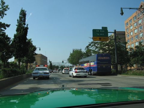 1991 Ford LTD Crown Victoria - Police cars trail a blue STARR charter bus on a city highway beneath a green 10 Ave exit sign, shot over a green car hood.